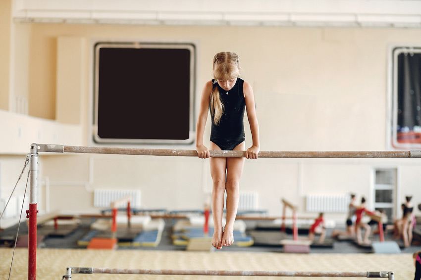 Κορίτσι με μαύρο κορμάκι γυμναστικής κρατιέται στους ασύμμετρους ζυγούς μέσα σε γυμναστήριο, συγκεντρωμένο στην προσπάθειά του. Η εικόνα αποτυπώνει τη στιγμή που το παιδί ξεπερνά τον φόβο του και περνάει από το «δεν μπορώ» στο «μπορώ».