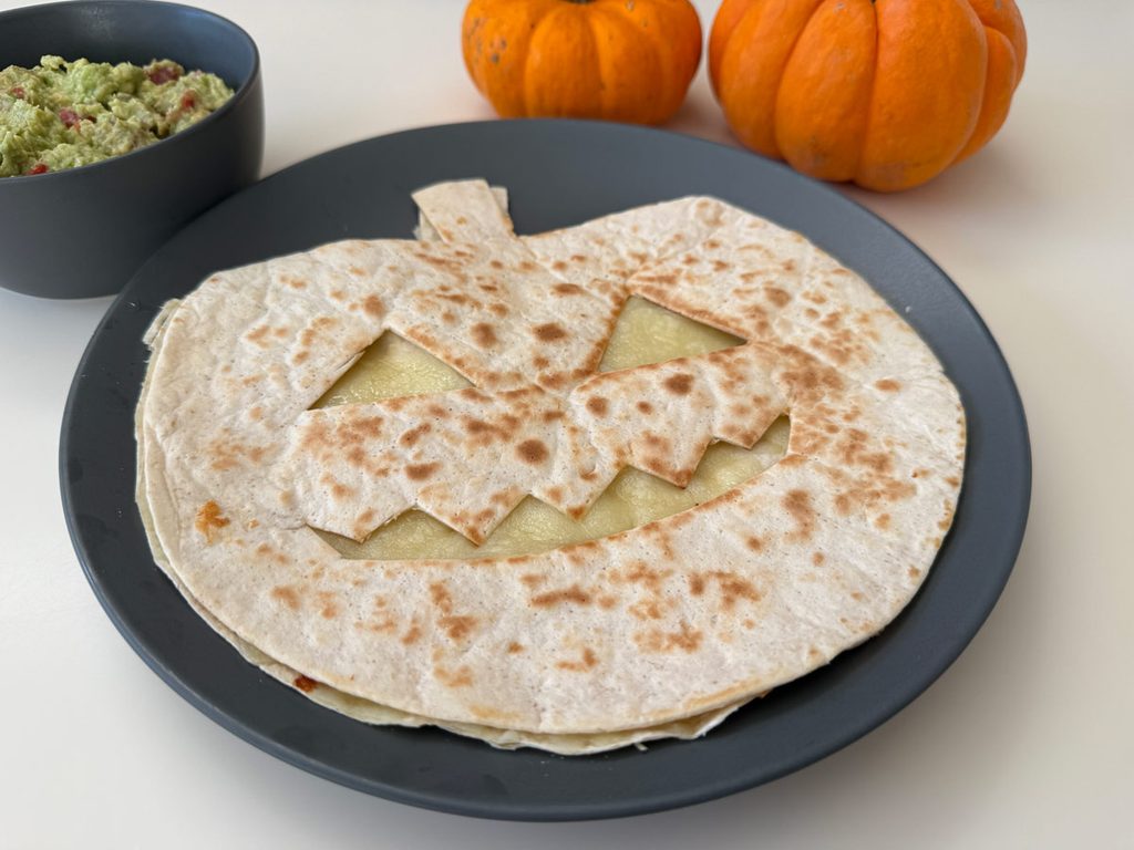 Έτοιμη quesadilla σε σχήμα κολοκύθας με χαμογελαστό πρόσωπο, από όπου φαίνεται το λιωμένο τυρί, σερβιρισμένη με γουακαμόλε και μικρές κολοκύθες δίπλα. Εύκολο φαγητό για Halloween με τα παιδιά.